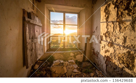 An empty room inside an old abandoned house featuring a stone wall, a dusty floor, and sunlight streaming through an ancient window near a dark fireplace in this urban architecture 136429688