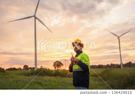 man in a yellow and black safety vest stands in front of two wind turbines 136429818