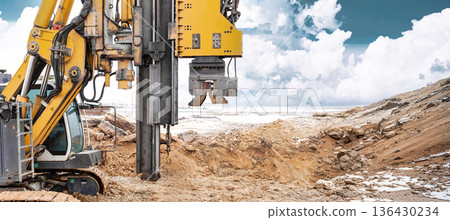 Drilling rig works on site to install piles into the ground. The area is active with dirt surrounding and clouds above. Work is ongoing in a construction zone Drilling rig works on site to install piles into the ground. The area is active with dirt surrounding and clouds above. Work is ongoing in a construction zone 136430234