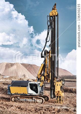 A drilling rig is set up at a construction site to install piles into the ground. The rig stands tall against a backdrop of clouds and earth mounds A drilling rig is set up at a construction site to install piles into the ground. The rig stands tall against a backdrop of clouds and earth mounds 136430235