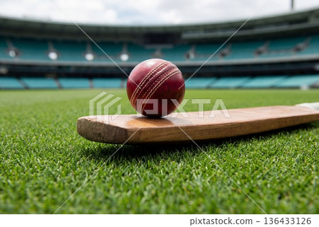 Red cricket ball balancing on a wooden bat on green grass in a large empty stadium field 136433126