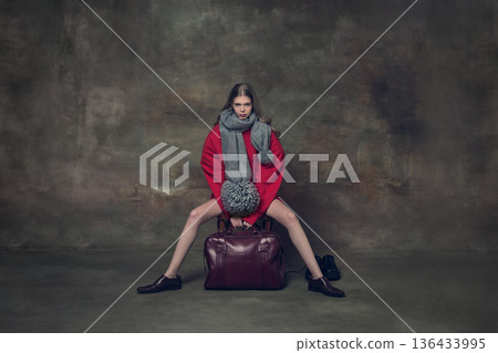 Young woman sitting wide legged on leather suitcase wearing oversized red sweater. 136433995