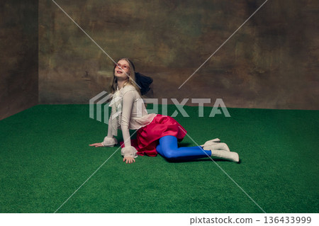 Young woman in colorful outfit lying on green studio floor smiling dreamily upward. 136433999