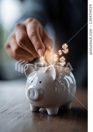 A man's hand drops a coin into a piggy bank on a table, and glowing dollar symbols appear around it, representing the concept of savings, personal finances, investments, and capital accumulation. 136434034