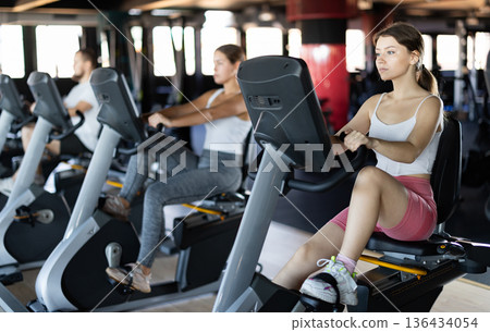 Girl performs exercises with stationary bicycle in gym Girl performs exercises with stationary bicycle in gym 136434054