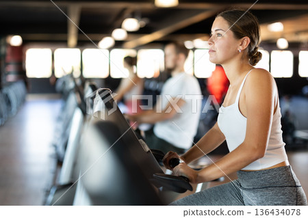 Girl performs exercises with stationary bicycle in gym 136434078
