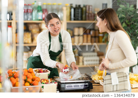 Woman sells and puts candies in bag for young woman, puts them on scale and weighs fruits Woman sells and puts candies in bag for young woman, puts them on scale and weighs fruits 136434079