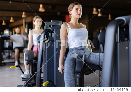 Slender athletic girl work out muscles of legs and thighs on simulator in fitness club Slender athletic girl work out muscles of legs and thighs on simulator in fitness club 136434159