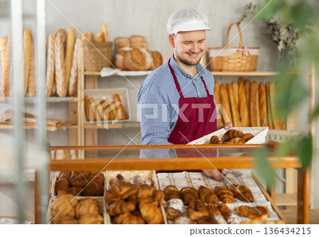 Man employee puts croissants in window, arranges display of goods at bakery. Man employee puts croissants in window, arranges display of goods at bakery. 136434215