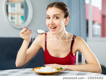 Young woman eating rice while sitting at the table in room. Diet and weight loss concept Young woman eating rice while sitting at the table in room. Diet and weight loss concept 136434262