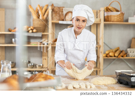 Skilled smiling young girl professional baker in white uniform standing at work table and kneading dough during working day in bakery 136434332