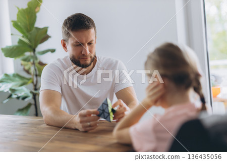 Father and daughter play with a puzzle cube at home, sitting at a wooden table and enjoying mindful time together 136435056
