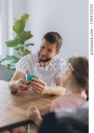 Father and daughter play with a puzzle cube at home, sitting at a wooden table and enjoying mindful time together Father and daughter play with a puzzle cube at home, sitting at a wooden table and enjoying mindful time together 136435059