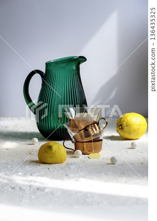 Emerald green glass jug and yellow lemon on white table against green wall.  136435325