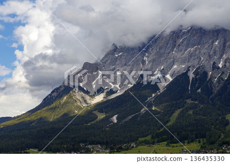 Scenic landscape view of Zugspitze mountain peak with clouds and green valley village. 136435330