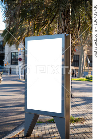 Blank outdoor advertising billboard mockup on a city street, providing copy space for promotion and marketing campaigns under warm sunlight next to a palm tree 136435430