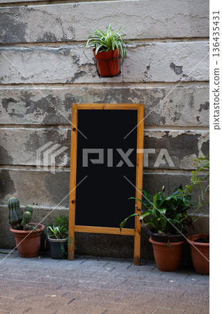 Blank chalkboard sandwich board with wooden frame standing on a paved sidewalk, surrounded by potted plants, against a textured weathered wall, offering space for advertising or menu display Blank chalkboard sandwich board with wooden frame standing on a paved sidewalk, surrounded by potted plants, against a textured weathered wall, offering space for advertising or menu display 136435431