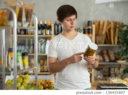 Young man store visitor chooses glass jar of pickled olives, hold and view glass jar package. Young man store visitor chooses glass jar of pickled olives, hold and view glass jar package. 136435807