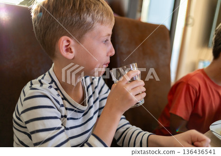 Elementary age boy drinking water at the table indoors, casual daily life moment Elementary age boy drinking water at the table indoors, casual daily life moment 136436541