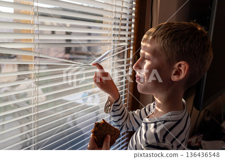 Boy looking through window blinds while holding a piece of cake, natural sunlight, indoor childhood lifestyle 136436548