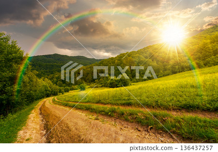 country road down the hill through the field at sunset. lovely countryside scenery of mountainous area before the summer storm in evening light. remote alpine pathway in rural highland under rainbow country road down the hill through the field at sunset. lovely countryside scenery of mountainous area before the summer storm in evening light. remote alpine pathway in rural highland under rainbow 136437257