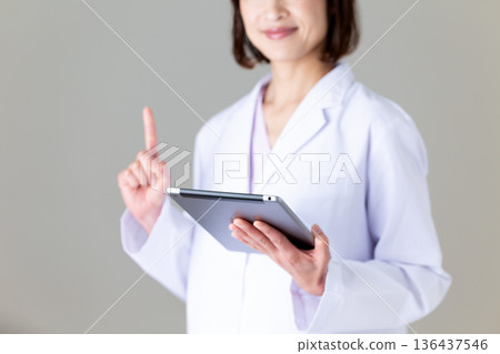 A middle-aged female nurse in a white coat holding a tablet and pointing her index finger 136437546