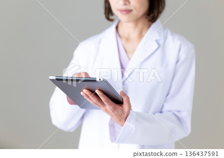 A middle-aged female nurse in a white coat holding a tablet 136437591