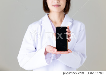 一位身穿白大褂的中年女護士，手裡拿著一支智慧型手機 136437780