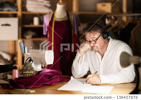 Middle-aged man drawing on paper in sewing workshop at night Middle-aged man drawing on paper in sewing workshop at night 136437878