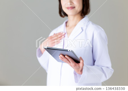 A middle-aged female doctor in a white coat holding a tablet and putting her hand on her chest 136438330