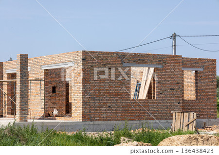 Builders work diligently on a brick house structure under clear skies, with tools and materials nearby in a serene setting Builders work diligently on a brick house structure under clear skies, with tools and materials nearby in a serene setting 136438423