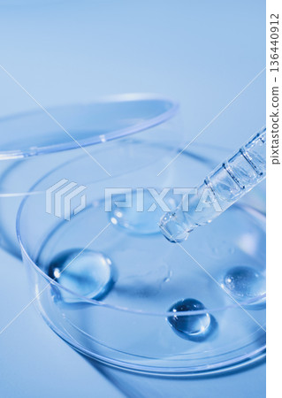 Pipette dropping clear liquid drops into clear petri dish on blue background Pipette dropping clear liquid drops into clear petri dish on blue background 136440912