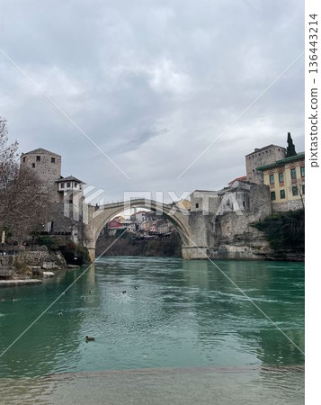 Front view of the Stari Most stone bridge spanning the Neretva river in Mostar with fortified towers and calm water under overcast sky Front view of the Stari Most stone bridge spanning the Neretva river in Mostar with fortified towers and calm water under overcast sky 136443214