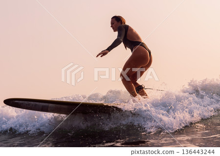 Attractive surfer woman in swimsuit rides on surfboard with warm sunlight. Woman on longboard on wave. Attractive surfer woman in swimsuit rides on surfboard with warm sunlight. Woman on longboard on wave. 136443244
