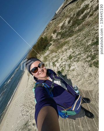 Solo female hiker taking selfie on sandy coastal trail along Atlantic ocean, Camino de Santiago in Portugal. Travel marketing, solo travel branding, outdoor lifestyle campaigns, adventure tourism 136445190