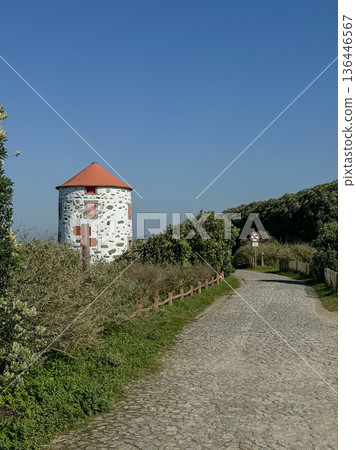 Cobblestone coastal path leading toward stone windmill with red roof along Camino de Santiago route in Portugal. Travel, pilgrimage route promotion, outdoor tourism communication, destination 136446567