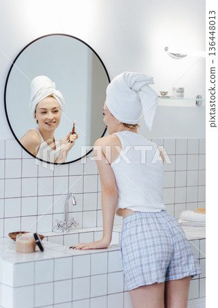 Blonde with a white towel on her head brushes her teeth in front of a mirror in a white bathroom. Blonde with a white towel on her head brushes her teeth in front of a mirror in a white bathroom. 136448013