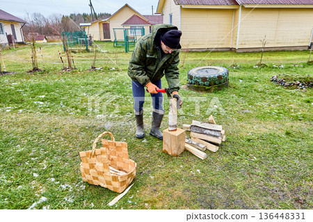 Splitting logs with axe into small pieces for lighting fire, mature man prepares kindling in his yard. 136448331