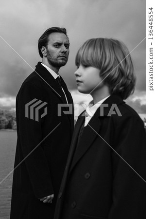 Serious father and daughter in dark coats outdoors under cloudy sky, black and white Serious father and daughter in dark coats outdoors under cloudy sky, black and white 136448554