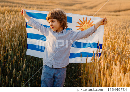 Patriot Boy Standing In Wheat Field Holding Uruguayan Flag In The Wind, Uruguay Patriot Boy Standing In Wheat Field Holding Uruguayan Flag In The Wind, Uruguay 136449784