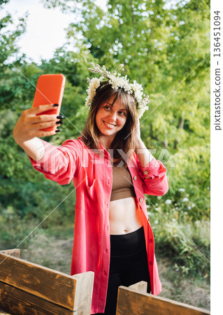 Smiling woman in flower crown taking selfie outdoors, summer festival vibe in green nature Smiling woman in flower crown taking selfie outdoors, summer festival vibe in green nature 136451094