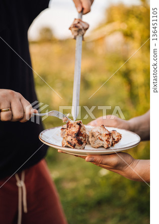 Serving grilled pork shashlik from skewer onto plate at outdoor barbecue 136451105