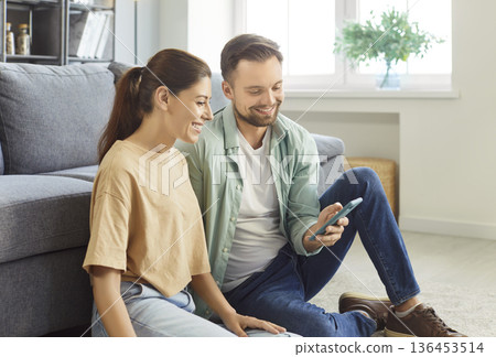 Family couple sitting comfortably on floor in modern living room smiling sharing mobile phone screen Family couple sitting comfortably on floor in modern living room smiling sharing mobile phone screen 136453514