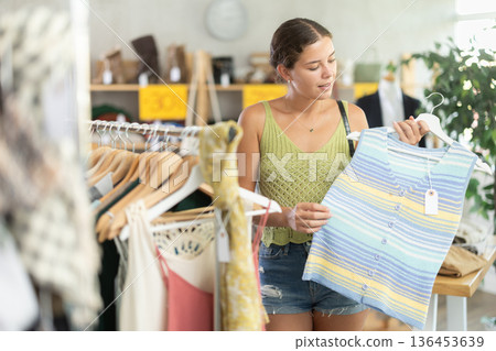 Positive young woman choosing something for summer in clothing store Positive young woman choosing something for summer in clothing store 136453639