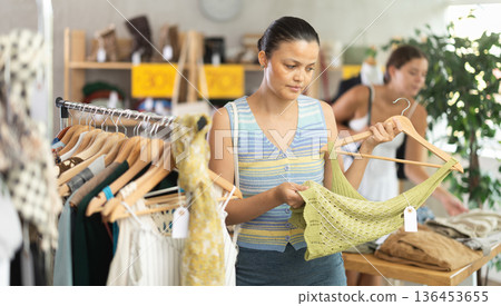Attentive young Asian woman choosing something for summer in clothing store Attentive young Asian woman choosing something for summer in clothing store 136453655