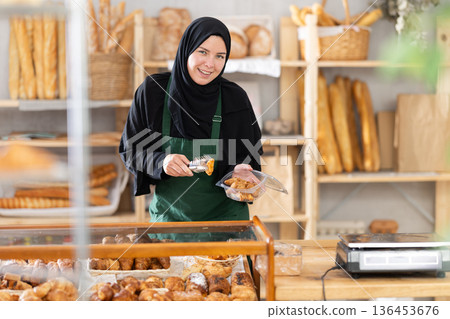Saleswoman in a hijab collects pastries in a container Saleswoman in a hijab collects pastries in a container 136453676