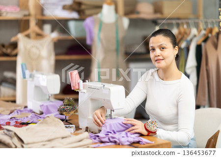 Attentive woman tailor sewing textile using sewing machine sitting at workplace in fashionable sewing atelier Attentive woman tailor sewing textile using sewing machine sitting at workplace in fashionable sewing atelier 136453677