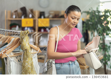 Near rack, Asian woman buys lady handbag, choose some items. 136453681