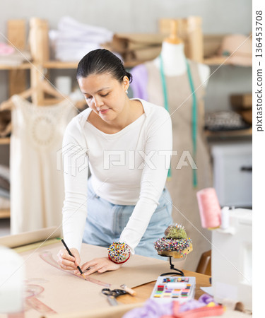 Young female seamstress drawing pattern on sheet of paper using curves while sewing dress at workplace in fashion sewing workshop Young female seamstress drawing pattern on sheet of paper using curves while sewing dress at workplace in fashion sewing workshop 136453708