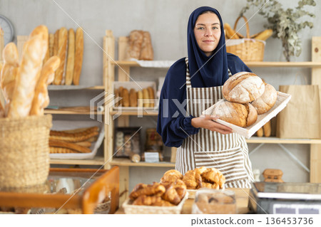 Muslim saleswoman holding loaves of bread in cozy bakery Muslim saleswoman holding loaves of bread in cozy bakery 136453736